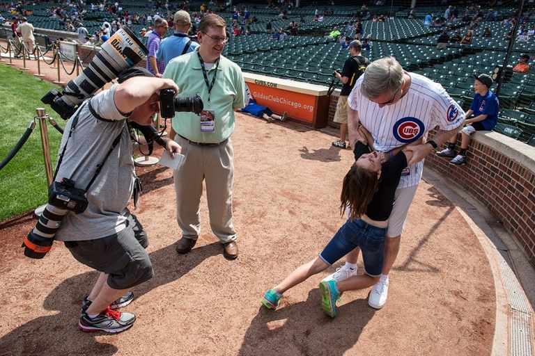 A photographer takes a photo of a man in a Cubs jersey playing with his 9 year-old daughter on a baseball field.