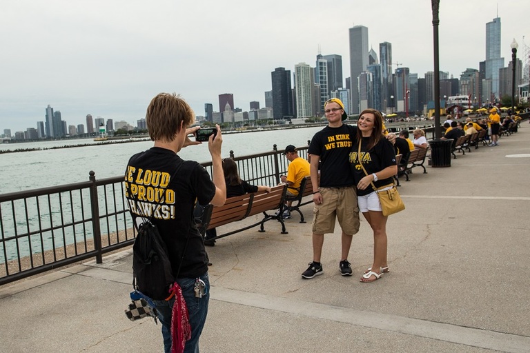 A man in a black t-shirt takes a photo of a couple on a pier with the Chicago skyline in the background.