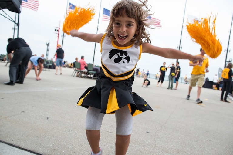 A 3 year old girl in a Hawkeye cheerleader uniform jumps in the air with pom-poms.