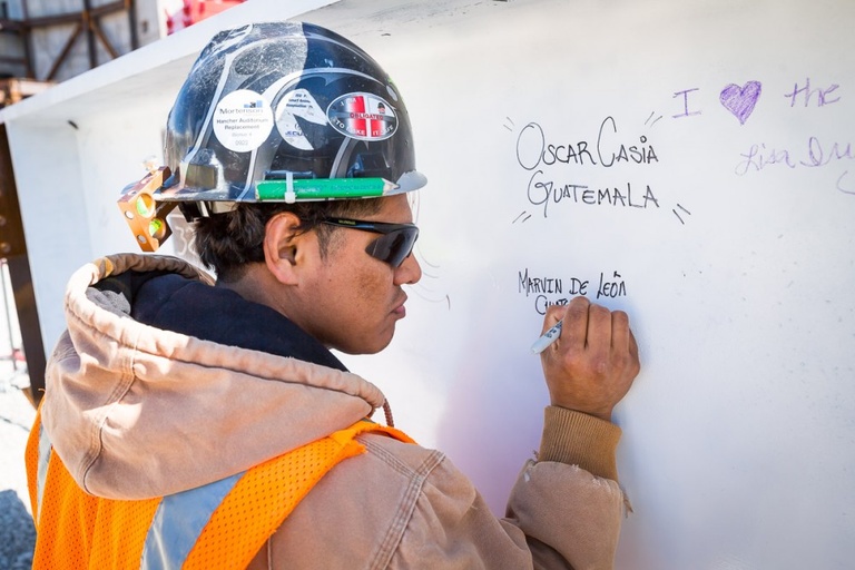 A man in a hard hat writes his name on a white wall with a marker.
