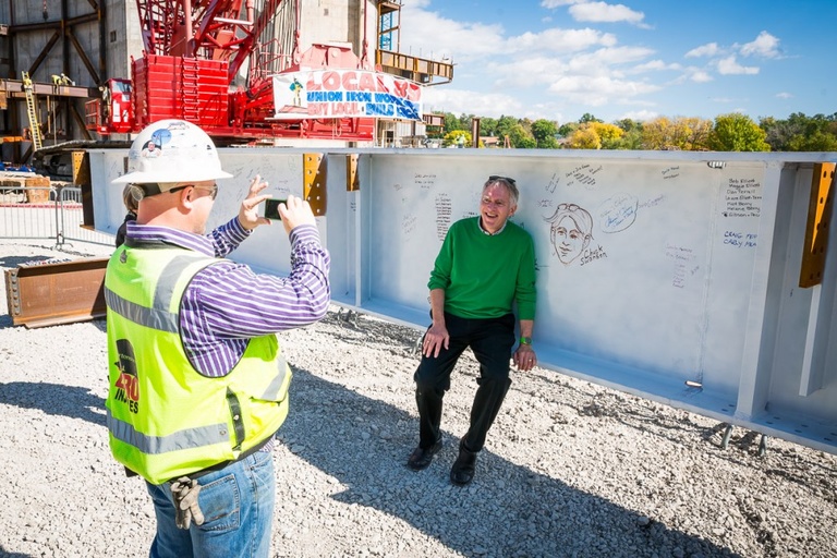 A man in a construction outfit takes a photo of a man in a green sweater.
