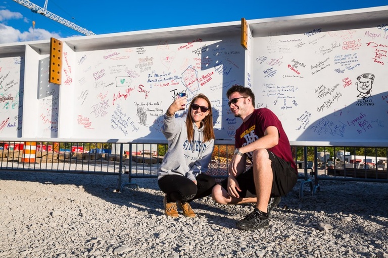 A young couple takes a selfie in front of a large white metal beam with signatures on it