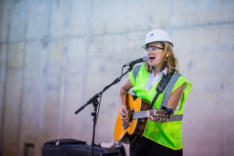 A woman in construction safety gear playing an acoustic guitar and singing into a microphone.