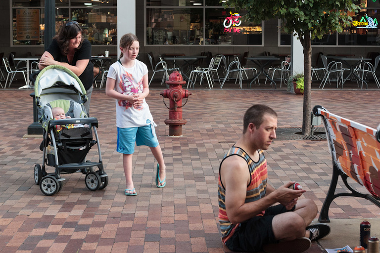 A woman pushing a stroller and a young girl observe a man spray painting a bench.