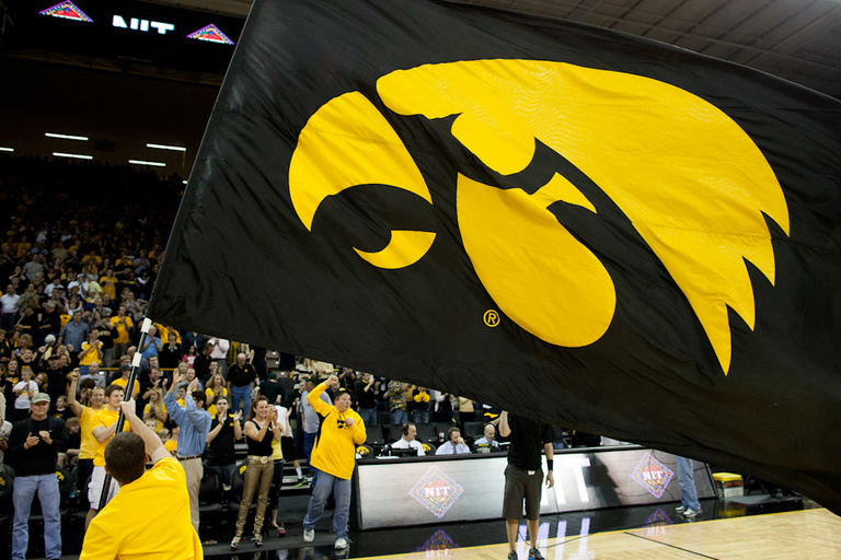An Iowa male cheerleader in the bottom left wearing yellow waves a large Tigerhawk flag on the floor of Carver-Hawkeye Arena while standing fans cheer in the backgound.