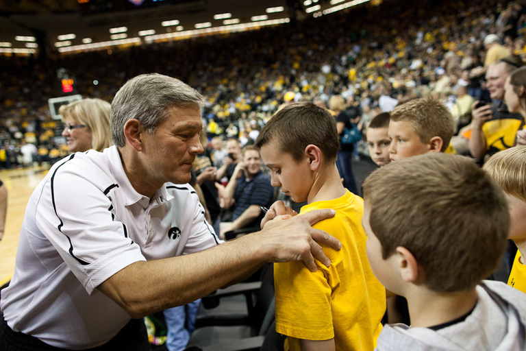 Iowa head football coach Kirk Ferentz autographs the yellow t-shirt of a young Iowa fan at courtside in Carver-Hawkeye Arena.