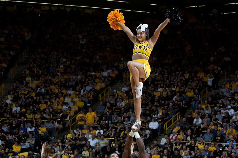An Iowa cheerleader balances on one foot held by a male cheerleader below her with her arms in the air and a full crowd at Carver-Hawkeye Arena behind her.
