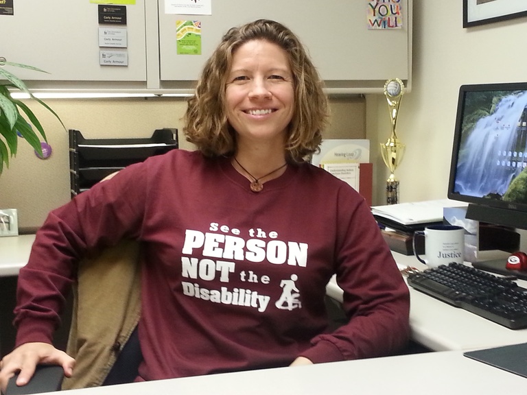 Carly Armour sits at her desk, wearing a "See the PERSON, NOT the disability" t-shirt