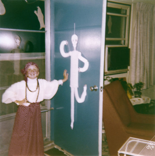 Color photo of young Jennifer Wilkins in costume, standing by an open door at Hawkeye Court Apartments on Halloween, circa 1978 