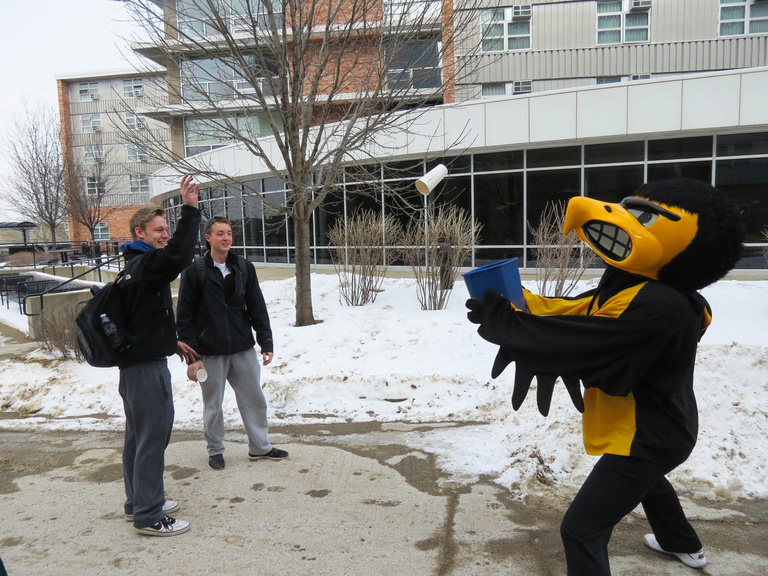 two students toss a paper cup into a recycle bin held by Herky