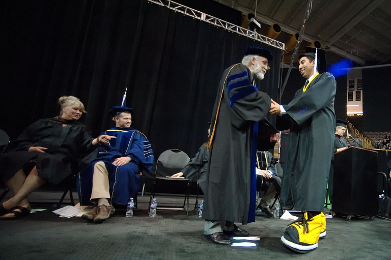 Graduating student shakes hand of Dean Djalali while wearing Herky's oversized sneakers.