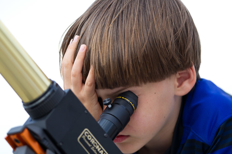 A young boy covers his right eye while looking into a telescope with his left.