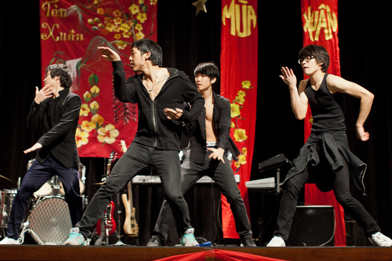 Four men wearing black pose mid-dance on stage with red Vietnamese draperies behind them.