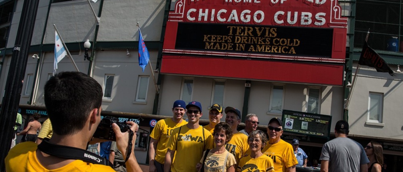 A man in a yellow t-shirt in the bottom left corner takes a photo of a group of people wearing Iowa Hawkeye and Cubs apparel in front of the Wrigley Field marquee.