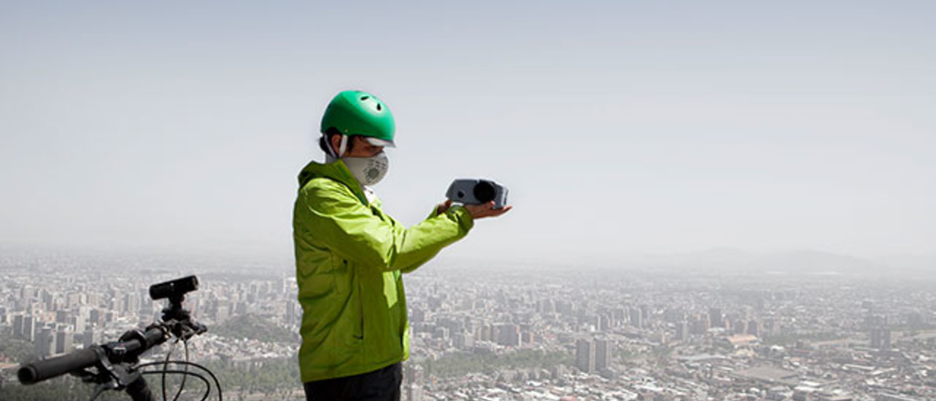Marcelo Mena-Carrasco is shown standing atop a hill overlooking Santiago with a portable monitoring device to measure levels of particulate matter in the air