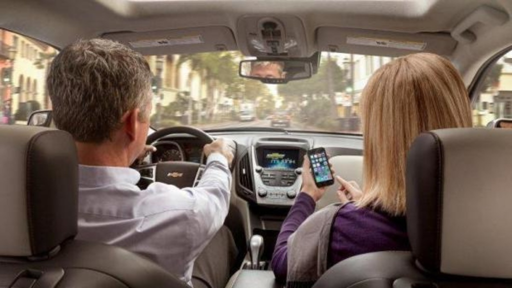 A man and a woman sit in the front seat of a car while the woman looks at a mobile device
