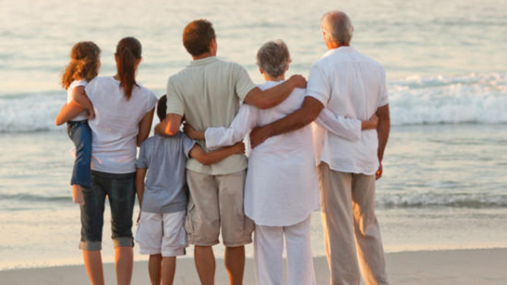 A photo of an intergenerational family standing on a beach looking out at the waves with their arms around each other.