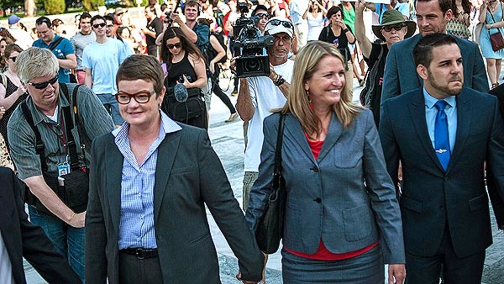 Kris Perry and Sandy Stier, two women, hold hands flanked by reporters and supporters after learning of the Supreme Court ruling that struck down portions of DOMA