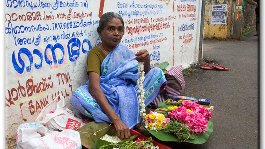 An image of a woman sitting on a street in India, surrounded by colorful flowers