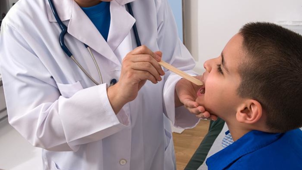 A pediatrician sticks a tounge depresser in a young boy&#039;s mouth as he shine a flashlight into his throat to check his tounsils