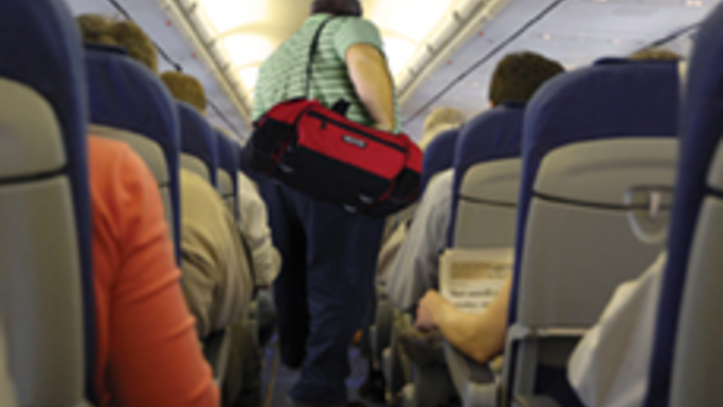 A large man carrying a red gym bag walks down an airplane aisle. 
