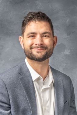 Casey DeRoo has short dark hair and a close cropped dark beard and mustache. He's wearing a light gray suitcoat and white dress shirt.