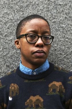Donika Kelly has short cropped dark hair, medium brown skin, dark round glasses and is wearing a navy blue sweater with brown bears, and a light blue dress shirt.