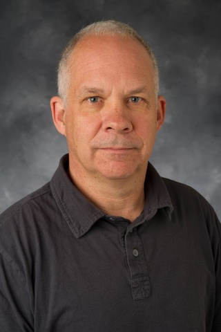 A close-up of Colin Gordon, a man with close cropped salt and pepper hair, wearing a black polo shirt.