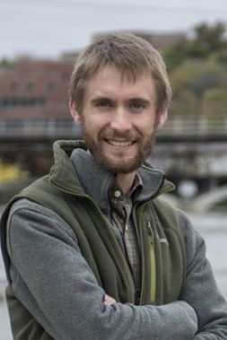 Greg LeFevre has sandy blonde hair, mustache and beard, is wearing a light gray sweater with a green fleece vest, and is standing along a river.
