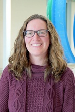 Colleen Bringman, with long, curly light brown hair, wearing glasses and a purple sweater
