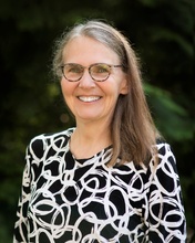 Polly Ferguson, a smiling woman, with medium length brown and gray hair. She's wearing tortoise shell patterned, round glasses and a black blouse with white swirls.