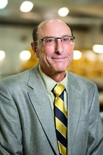 Fred Stern, a man wearing a blue green suit jacket and blue and yellow striped tie. He has dark hair and is wearing black, horned rim glasses.