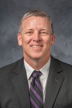 Ted Neal, smiling, with short cropped blond and gray hair, wearing a gray pinstriped suit jacket and purple striped tie.