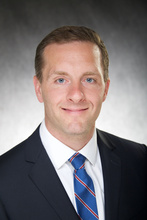 Chad Tracy, a man with short, reddish-blonde hair, bright blue eyes, wearing a dark suit jacket and blue and red striped tie.