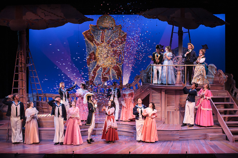 Far away shot of a theater stage set as a sailboat theme. A group of men and women dance together as colorful confetti falls from the ceiling.