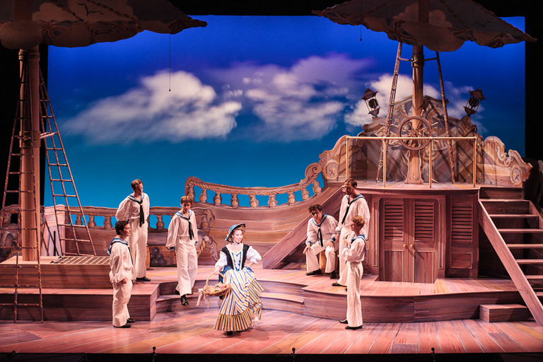 Far away shot of a theater stage set as a sailboat theme. Actors dressed as seamen surround a woman singing in a dress.
