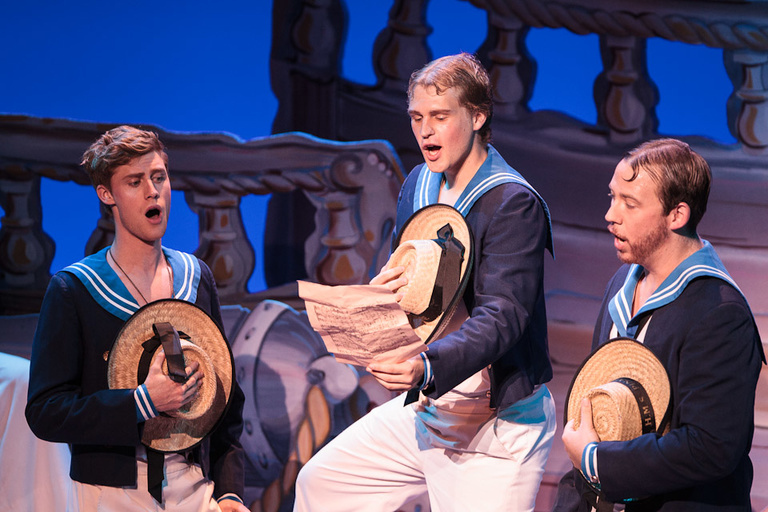 Three men dressed as sailors singing, the middle man holding a piece of parchment.