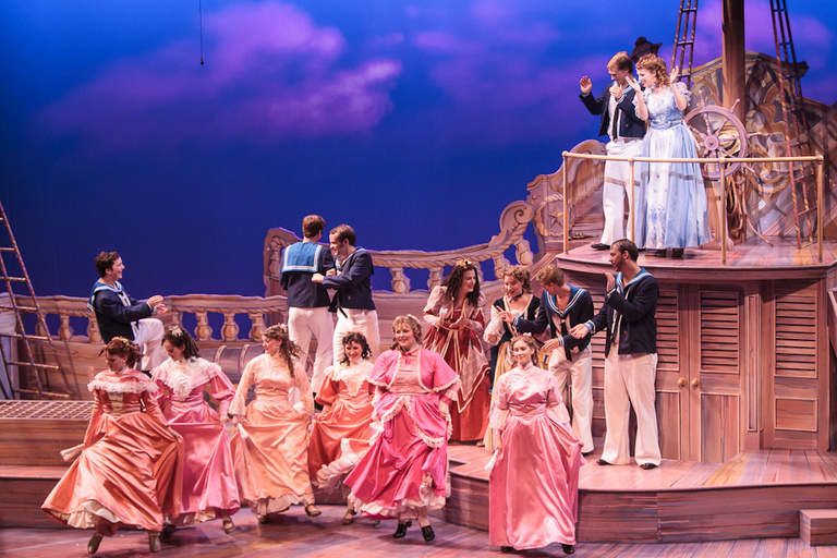 Far away shot of a theater stage set as a sailboat theme. A group of women dance in dresses in the foreground while men dressed as sailors dance behind them.