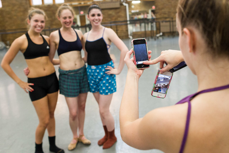 Three female dancers in the background pose for another female dancer taking their photo on an iPhone.