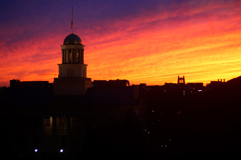A photo of the University of Iowa Old Capitol on the Pentacrest in silhouette as the sun sets with gold, red, and yellow streaks in the sky as darkness falls.