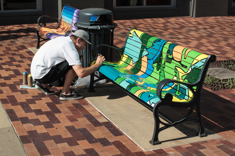 A man kneels in front of a painted bench, drawing on it with a black paint marker.
