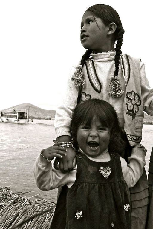 Image of two young Quechuan sisters who live on a floating island in Peru with the younger girl in front and with the older sister's hand on her shoulder with her face not visible.