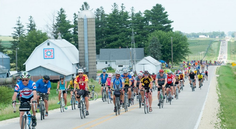 A stream of riders ride past a quilt barn.
