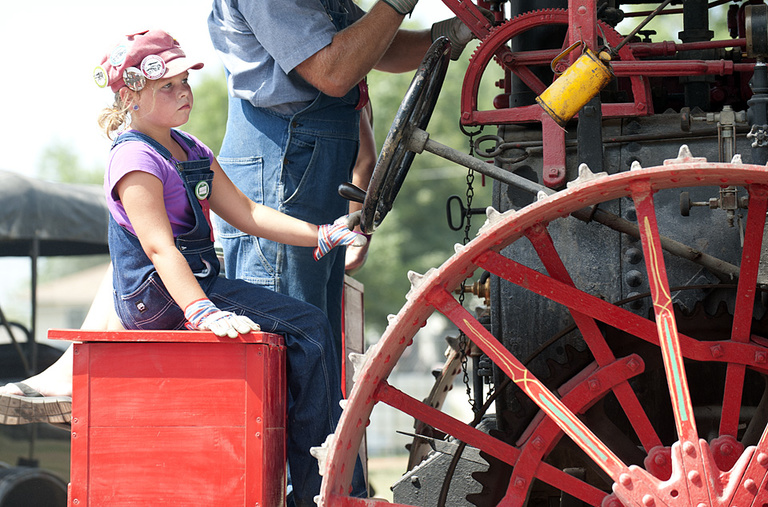 A young girl helps drive a big steam tractor.