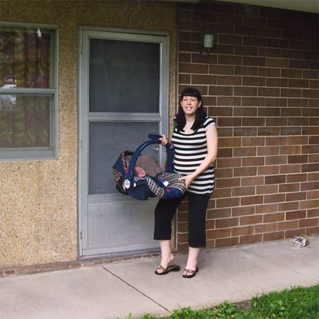 Color photo of Anne Wilmoth posing outside the door of her Hawkeye Court apartment holding her newborn baby in a carseat