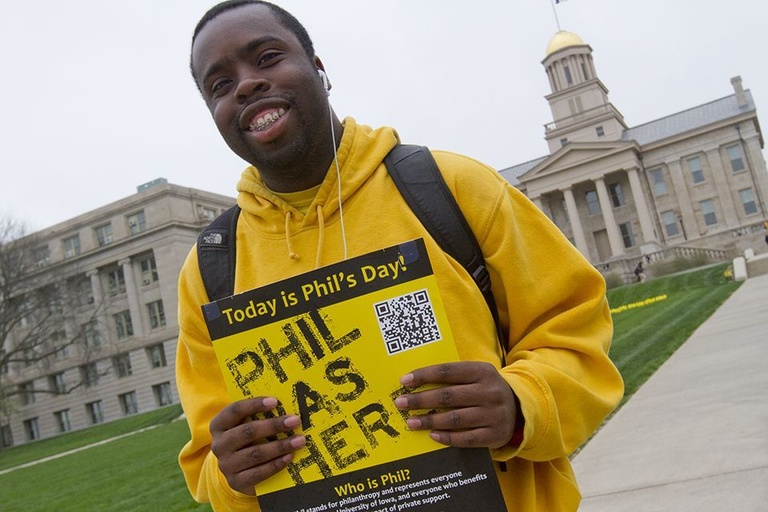 A student standing outside of the Old Capitol on the Pentacrest holds a sign announcing that "Phil was here."