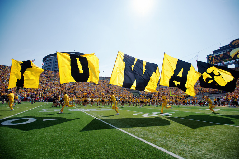 Cheerleaders run across football field with I-O-W-A flags.