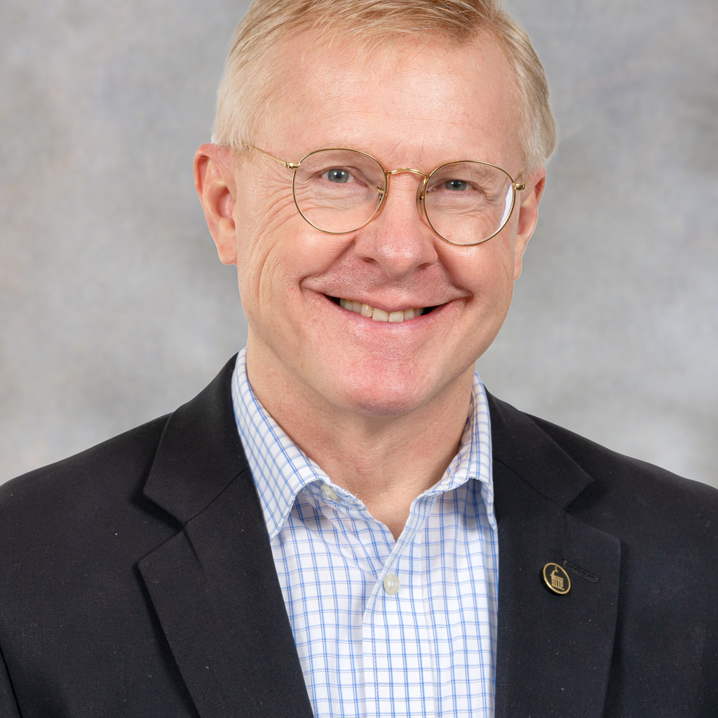 Nick Street, a man with strawberry-blond, short hair wearing round glasses. He's wearing a dark suit jacket and a blue checked shirt. 