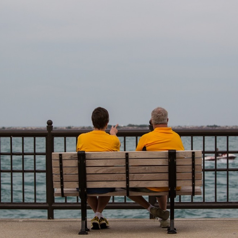 A couple in yellow t-shirts sit on a bench and look out over a body of water.