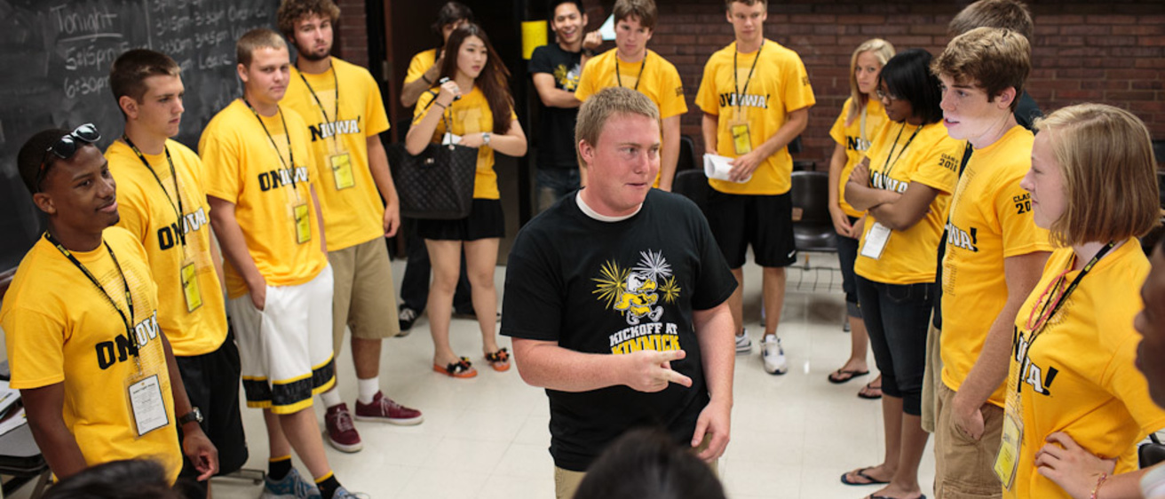 A man in a black t-shirt surrounded by people in yellow t-shirts in a classroom.